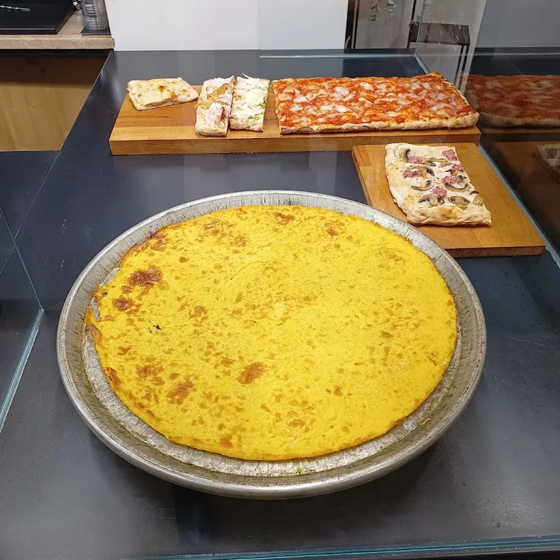 Una cecina rotonda e dorata nella sua teglia, accanto a taglieri con tranci di pizza.