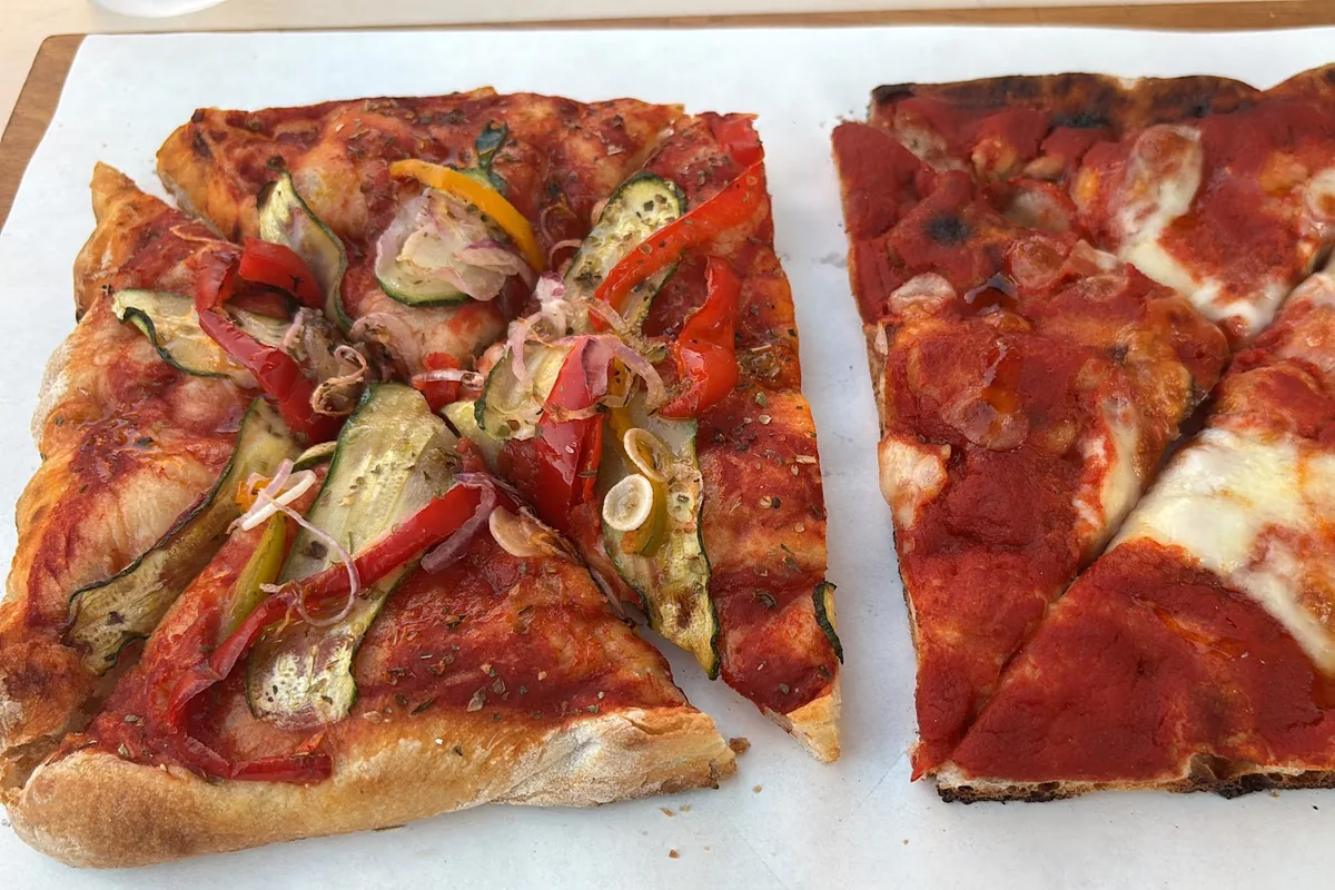 Teglie di pizza sulla carta, tagliate in tranci: la prima con verdure grigliate sul pomodoro.